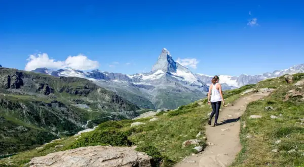 Frau wandert auf einem Pfad mit Blick auf das majestätische Matterhorn. Die Berglandschaft ist grün und felsig, der Himmel strahlend blau.