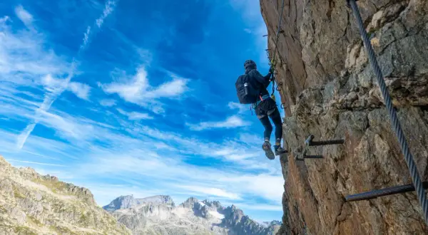 Ein Klettersteig in der Schweiz: Eine Person klettert gesichert an einem Felsen entlang, mit Blick auf majestätische Berge unter blauem Himmel.