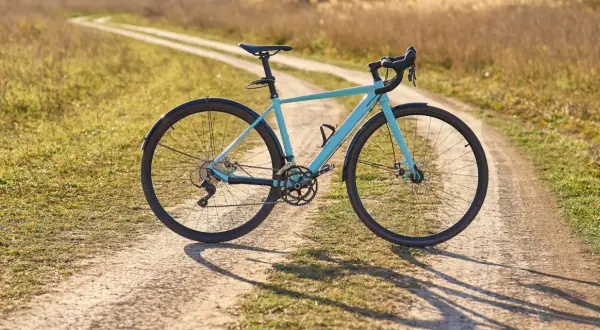 Ein hellblaues Fahrrad mit Schutzblechen steht auf einem Feldweg. Der Unterschied zwischen Cyclocross und Gravel wird hier deutlich: Dieses Rad ist bereit für Abenteuer auf Schotterwegen.
