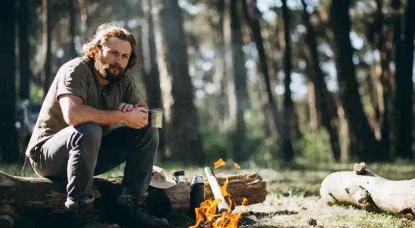 Mann sitzt am Lagerfeuer im Wald, hält eine Tasse. Das ist Bushcraft: Naturverbundenheit und Selbstversorgung.