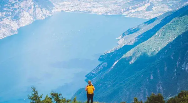 Frau mit gelbem Rucksack steht auf einem Berg und blickt auf einen See. Ein traumhafter Ausblick auf die italienischen Fernwanderwege.