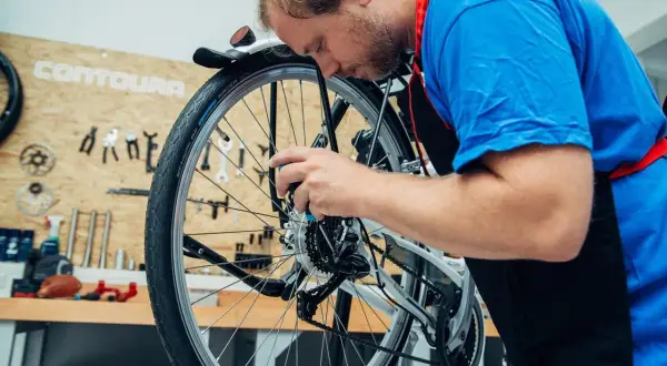 Ein Mann mit Bart im blauen T-Shirt und schwarzer Schürze arbeitet konzentriert am Fahrrad. Er wechselt das Fahrrad Schaltwerk.