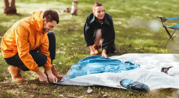 Mann und Frau bauen ein Zelt auf. Sie legen die Zeltplane aus und befestigen sie im Gras.