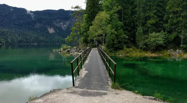 Ein schmaler Steg führt über das smaragdgrüne Wasser des Eibsees. Der eibsee rundweg lädt zu einer Erkundung der malerischen Natur ein, umgeben von dichten Wäldern und Bergen.