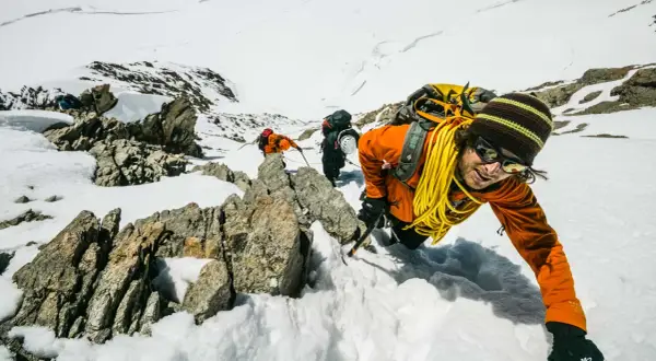 Bergsteiger kämpfen sich durch den Schnee in den Berner Alpen. Ein Mann in Orange lächelt in die Kamera, während er sich an einem Felsen festhält.