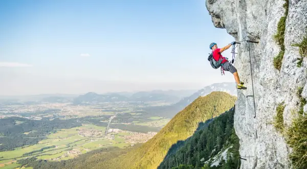 Ein Mann klettert gesichert an einem Stahlseil einen Felsvorsprung entlang. Der Klettersteig in Deutschland bietet eine atemberaubende Aussicht auf das Tal.