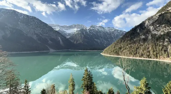 Spiegelglatter See am Plansee Panoramaweg, umrahmt von bewaldeten Bergen und schneebedeckten Gipfeln. Ein perfekter Tag für eine Wanderung.