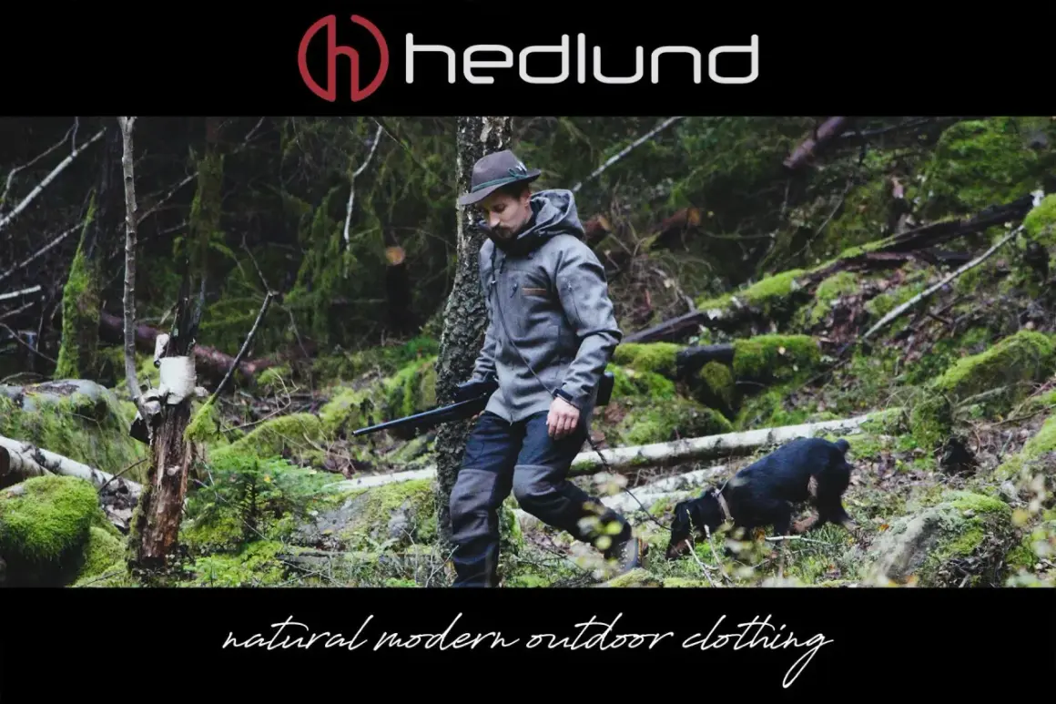 Ein Mann in Lodenkleidung und Hut geht mit einem Hund und einem Gewehr durch einen moosbewachsenen Wald.