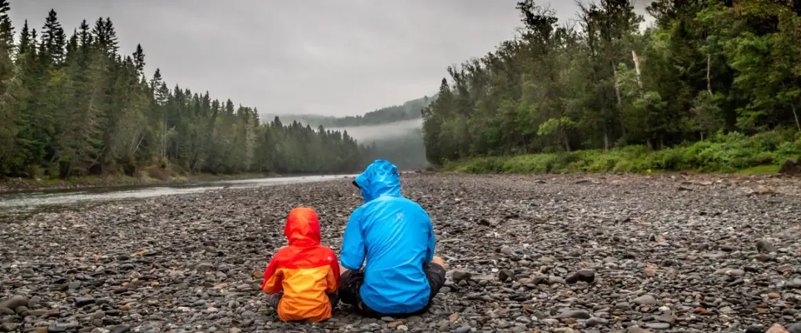 Zwei Personen in bunten Regenjacken sitzen am steinigen Ufer eines Flusses. Die richtige Gore-Tex-Pflege hält sie trocken.