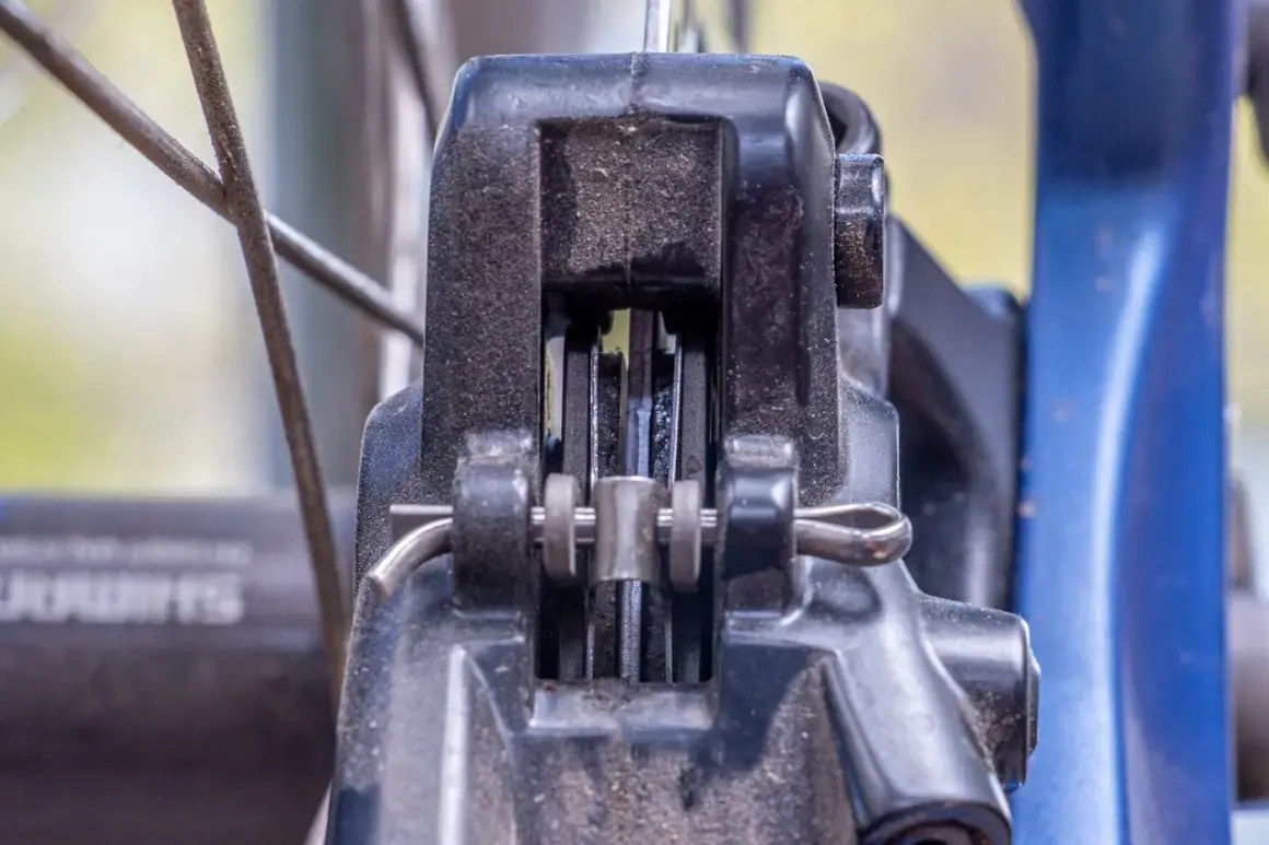 Nahaufnahme einer Fahrradbremse. Die Bremsbeläge schleifen einseitig am Rotor. Staub und Schmutz sind sichtbar.