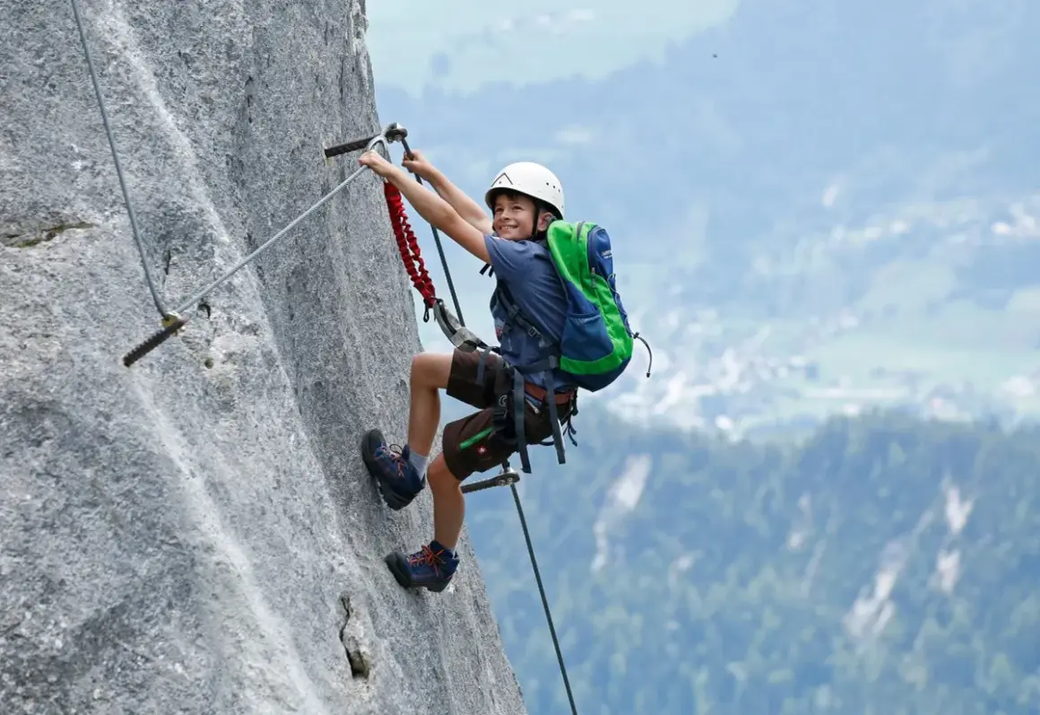 Ein Junge mit Rucksack und Helm meistert einen Klettersteig. Ein tolles Abenteuer für klettersteig kinder!