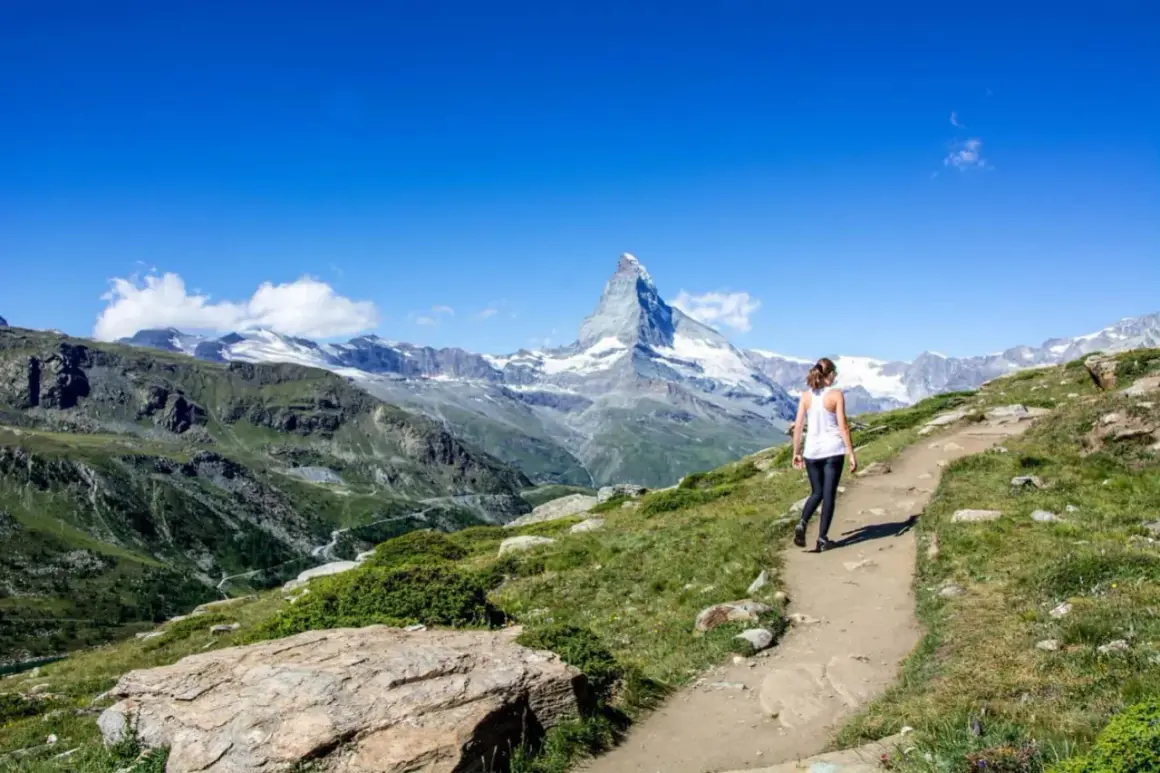 Frau wandert auf einem Pfad mit Blick auf das majestätische Matterhorn. Die Berglandschaft ist grün und felsig, der Himmel strahlend blau.