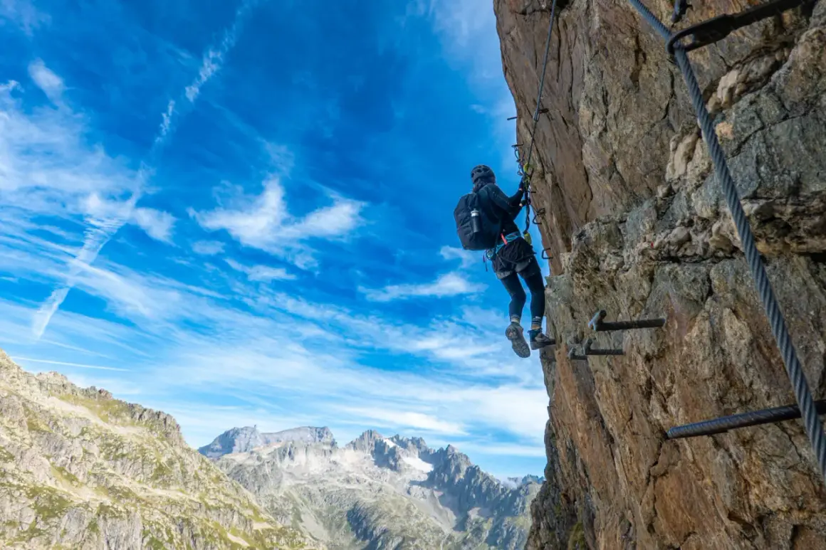 Ein Klettersteig in der Schweiz: Eine Person klettert gesichert an einem Felsen entlang, mit Blick auf majestätische Berge unter blauem Himmel.
