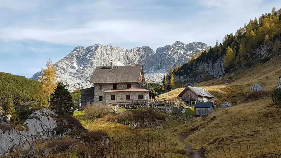 Steinhäuser und Holzhütten am Fuße eines majestätischen, schneebedeckten totes Gebirge. Herbstliche Bäume leuchten golden.