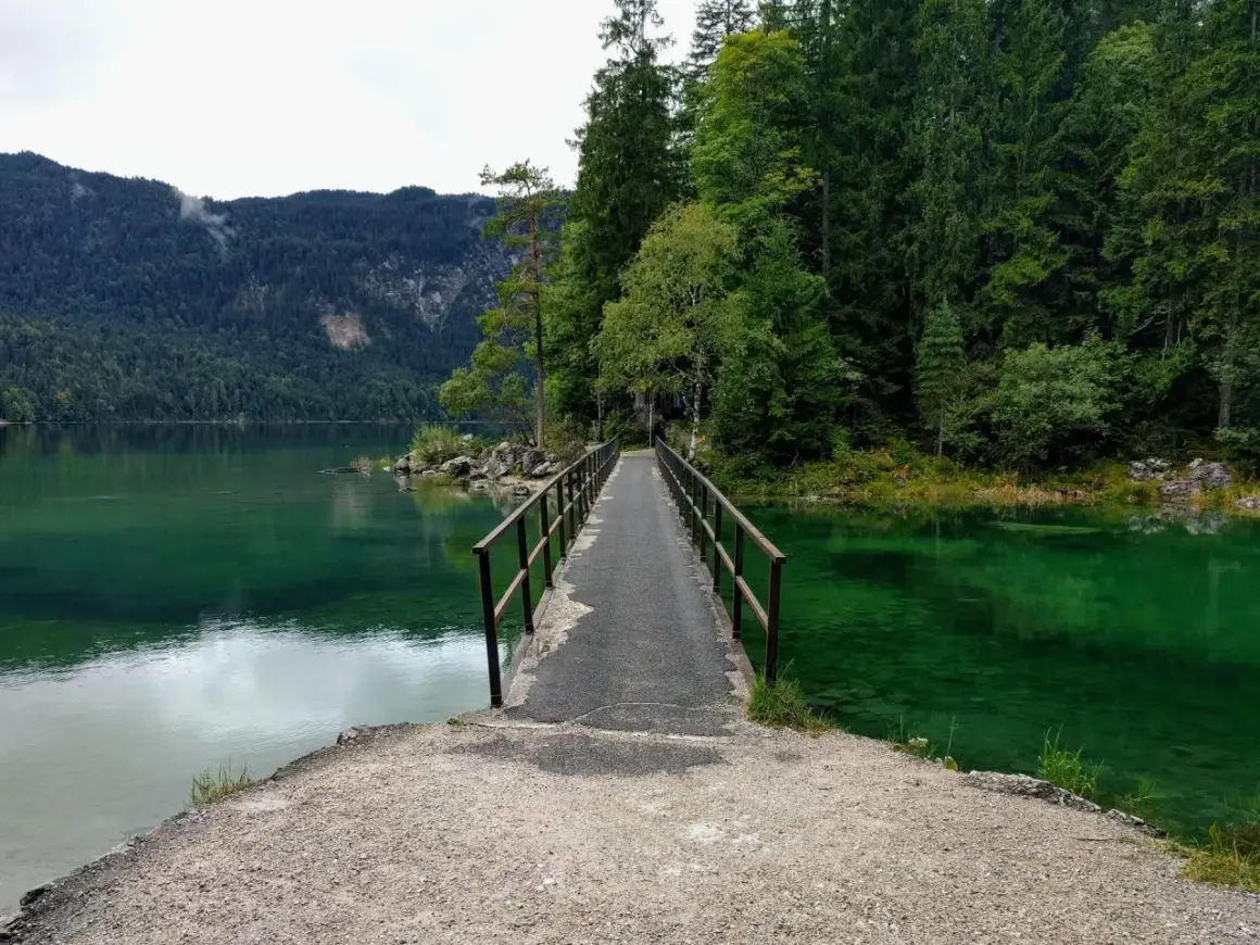 Ein schmaler Steg führt über das smaragdgrüne Wasser des Eibsees. Der eibsee rundweg lädt zu einer Erkundung der malerischen Natur ein, umgeben von dichten Wäldern und Bergen.