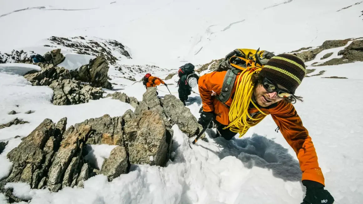 Bergsteiger kämpfen sich durch den Schnee in den Berner Alpen. Ein Mann in Orange lächelt in die Kamera, während er sich an einem Felsen festhält.