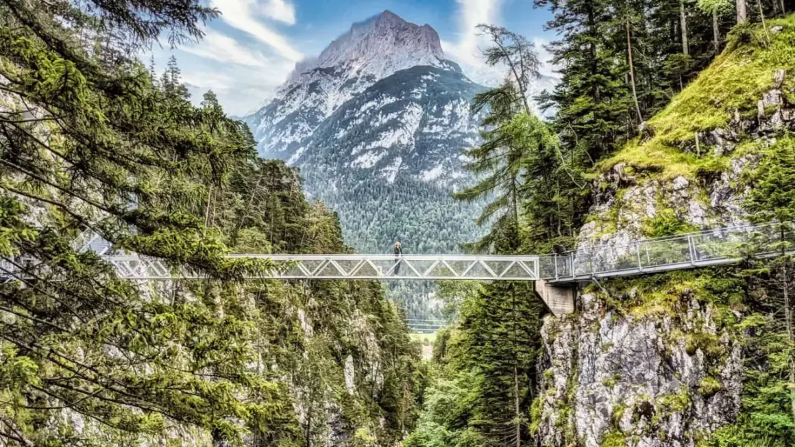 Ein Wanderer überquert eine Hängebrücke imalat, umgeben von grünen Wäldern und mit einem majestätischen Berg im Hintergrund.