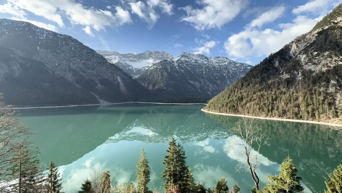 Spiegelglatter See am Plansee Panoramaweg, umrahmt von bewaldeten Bergen und schneebedeckten Gipfeln. Ein perfekter Tag für eine Wanderung.