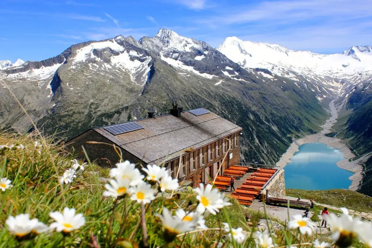 Alpenh&uuml;tte mit Solarpanels, davor eine Terrasse mit Tischen, im Hintergrund schneebedeckte Berge und ein blauer Bergsee. Ein perfekter Ort f&uuml;r die Peter Habeler Runde.
