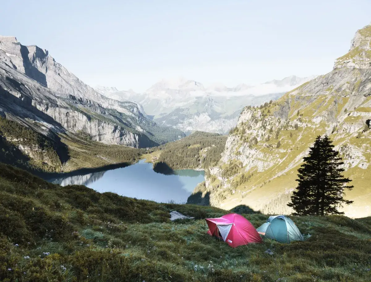 Zwei Zelte, ein rotes und ein gr&uuml;nes, stehen auf einer gr&uuml;nen Wiese mit Blick auf einen Bergsee. Ein perfekter Ort f&uuml;r wildcampen in Europa.
