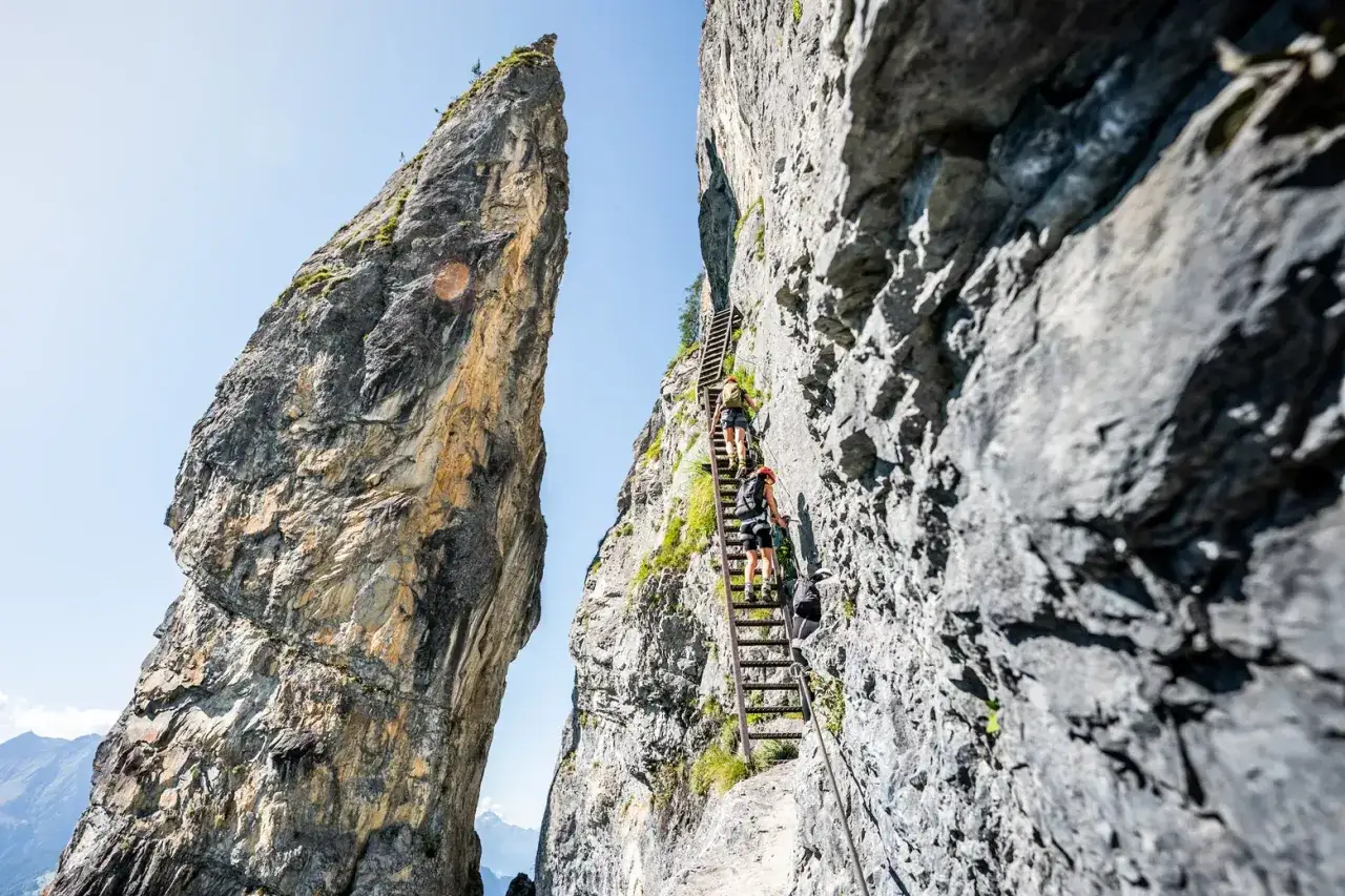 Zwei Wanderer erklimmen eine steile Leiter an einem Felsvorsprung. Ein atemberaubender Klettersteig in der Schweiz, der Abenteuer und spektakuläre Ausblicke verspricht.