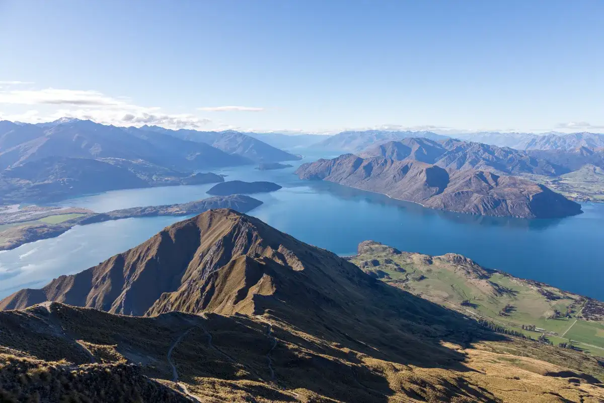 Atemberaubender Blick vom Roys Peak auf den tiefblauen See und die umliegenden Berge. Ein Pfad schlängelt sich den Hang hinauf.