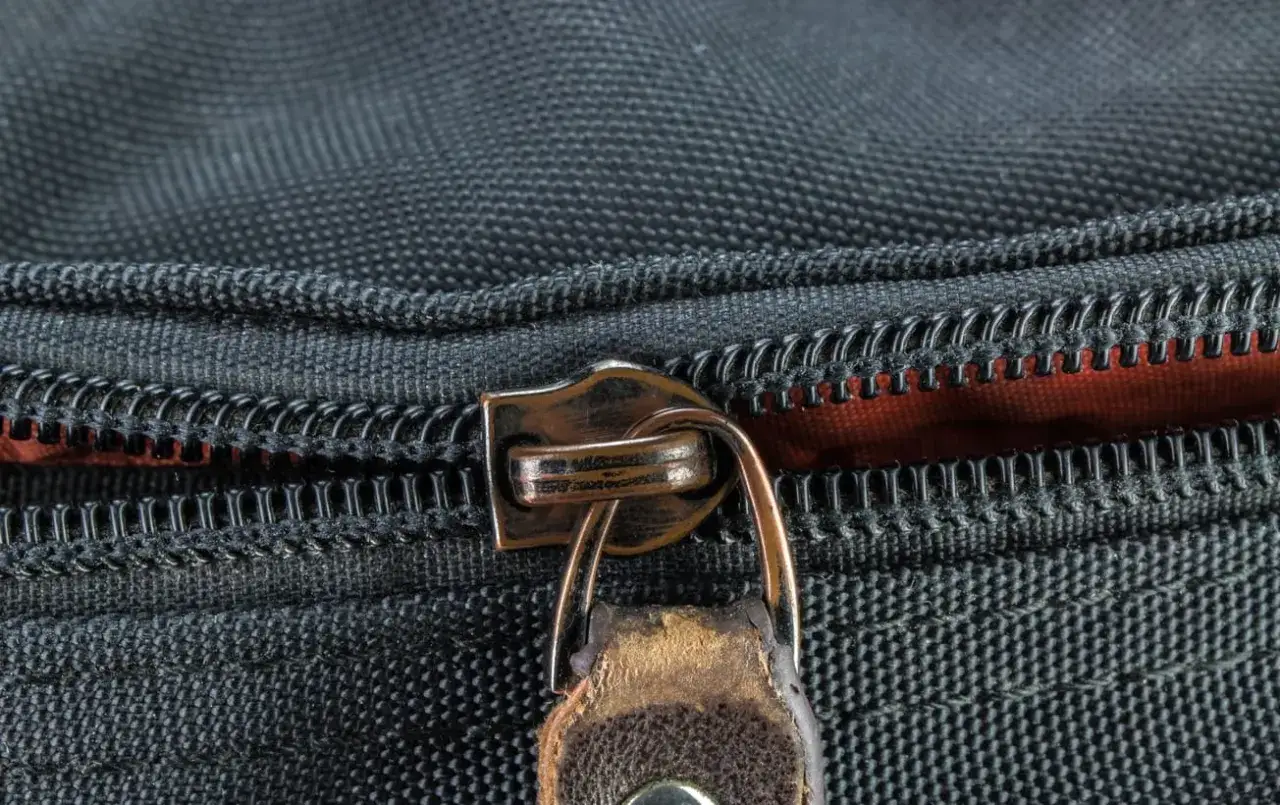 Nahaufnahme eines Rei&szlig;verschlusses an einer schwarzen Tasche. Der Zipper ist aus Metall und leicht abgenutzt. Ein kleiner Lederanh&auml;nger ist daran befestigt. Hier k&ouml;nnte man den Rei&szlig;verschluss reparieren.