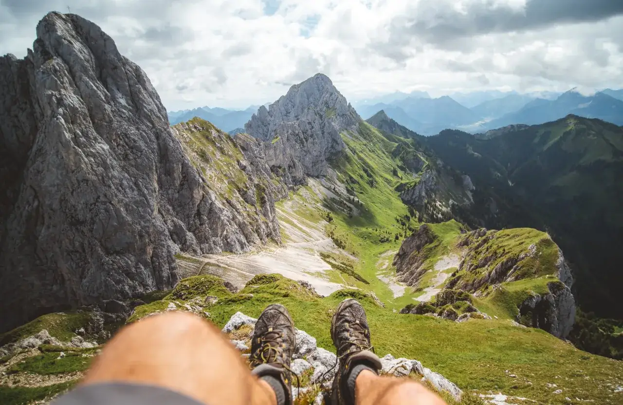 F&uuml;&szlig;e in Wanderschuhen auf einem Berggipfel im Tannheimer Tal. Gr&uuml;ne H&auml;nge und felsige Gipfel erstrecken sich in die Ferne.