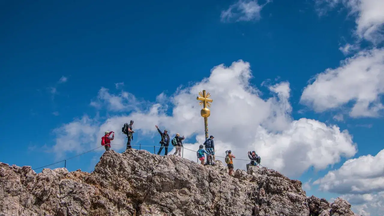 Menschen auf dem felsigen zugspitze gipfel unter blauem Himmel mit goldenem Kreuz und Wolken.