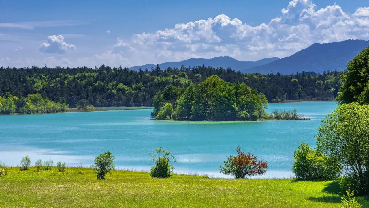 Türkisfarbener See, grüne Wiesen und ein Wald laden zu einer leichten Wanderung ein. Berge im Hintergrund und eine kleine Insel im See.