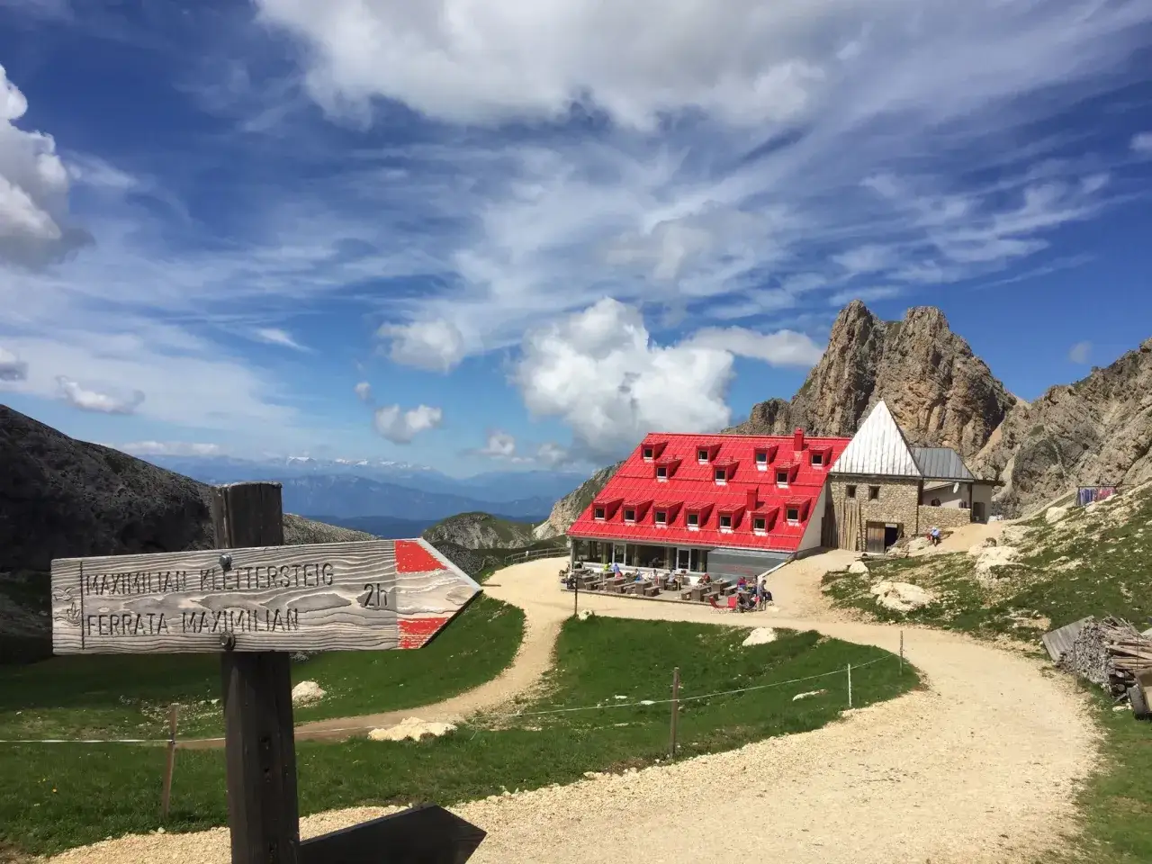 Wanderwegweiser zu den Maximilian Klettersteig und Ferrata Maximilian. Einladende Hütte mit roten Dach, ideal für fernwanderwege Italien.
