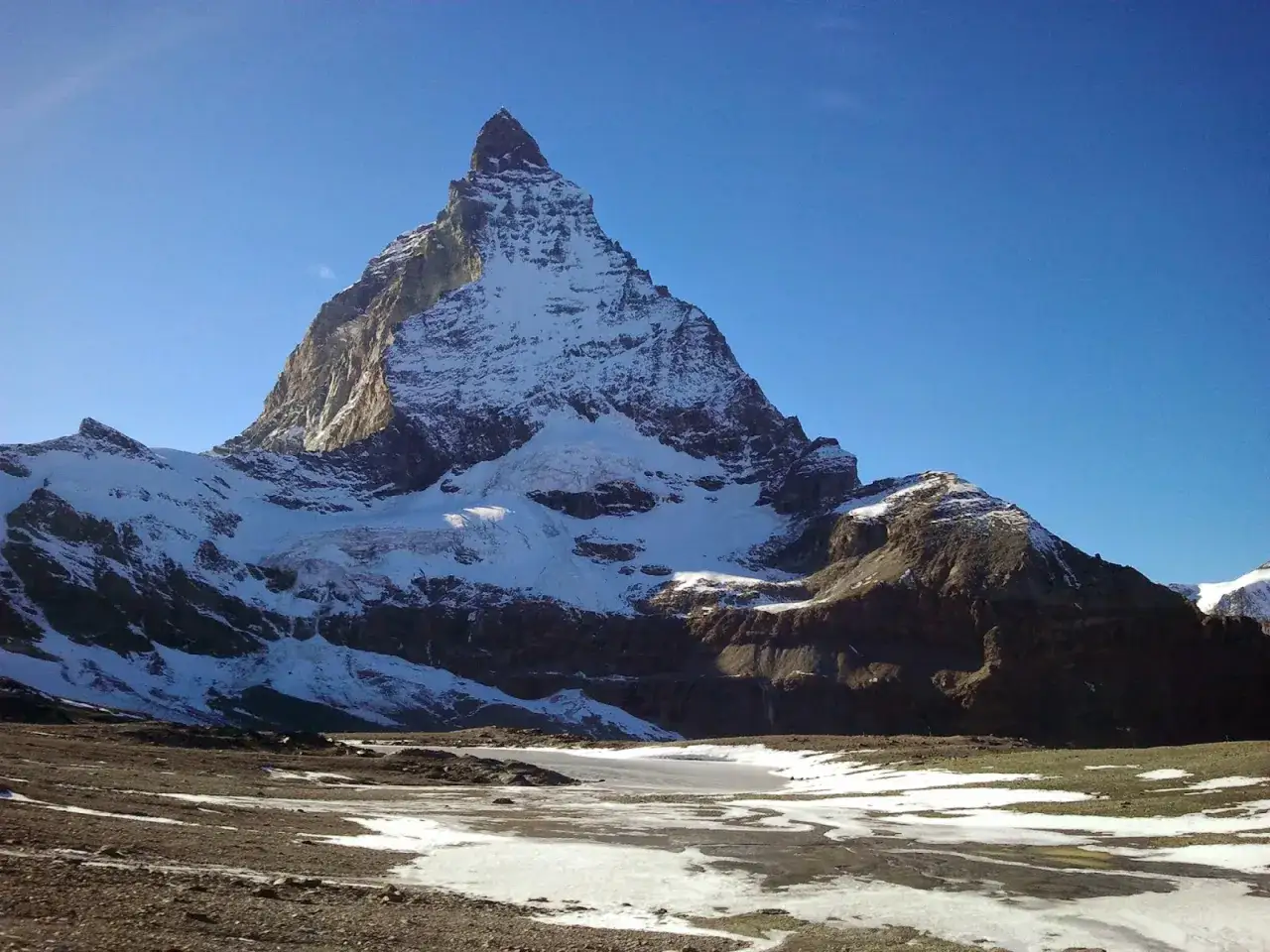 Das Matterhorn mit seiner imposanten Nordwand ragt schneebedeckt in den blauen Himmel. Vor dem Berg liegt eine eisbedeckte Fläche.