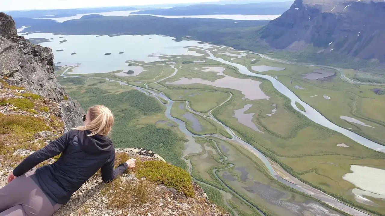 Blick von einem Felsen auf eine weite Flusslandschaft, die an eine der Kungsleden Etappen erinnert. Grüne Wiesen und Wälder durchzogen von schimmernden Wasserläufen.