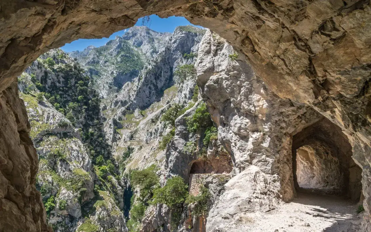 Blick durch einen Felsbogen auf die zerklüfteten Berge der Picos de Europa. Ein schmaler Pfad führt in einen dunklen Tunnel.