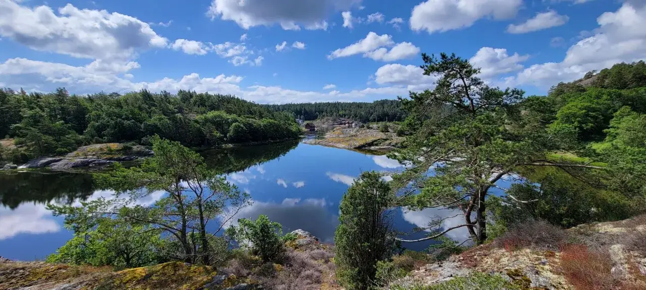 Idyllische Bucht mit roten Häusern, perfekt für Schweden-Wandern. Spiegelglattes Wasser reflektiert Wolken und grüne Wälder.