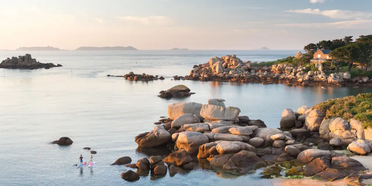 Zwei Personen paddeln auf Stand-Up-Boards im ruhigen Wasser vor einer felsigen K&uuml;ste der Bretagne. Ein Haus thront auf einem H&uuml;gel.