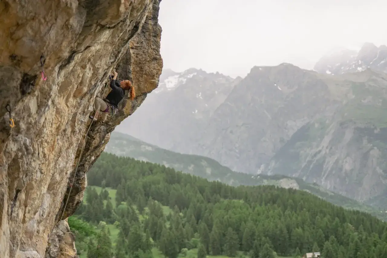 Frau mit roten Haaren klettert an einer Felswand bei Brian&ccedil;on. Im Hintergrund erheben sich bewaldete Berge und schneebedeckte Gipfel.