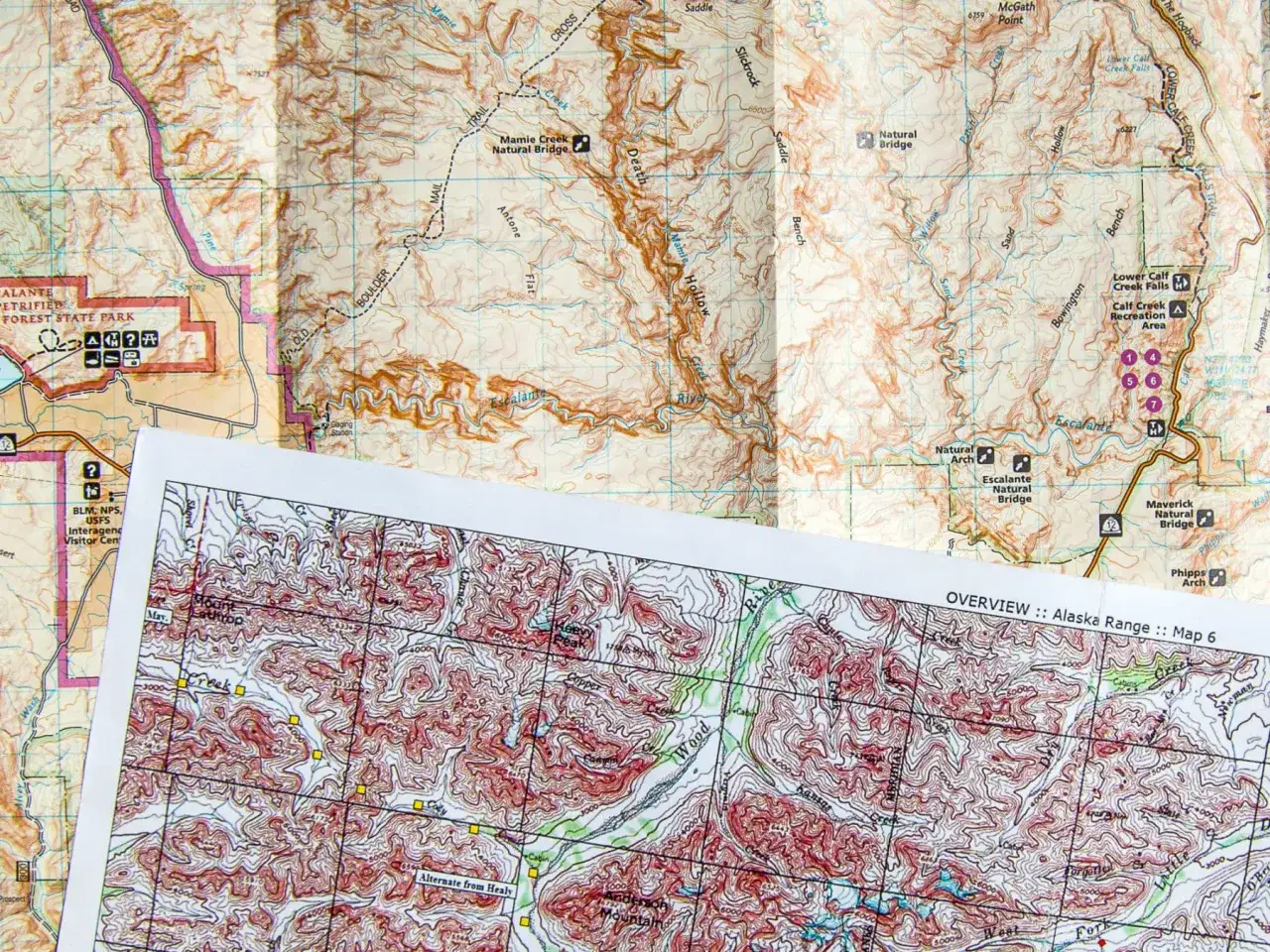 Zwei detaillierte topografische Karten liegen übereinander. Eine davon zeigt den Escalante River und die Alaska Range. Perfekt, um eine Wanderung zu planen.