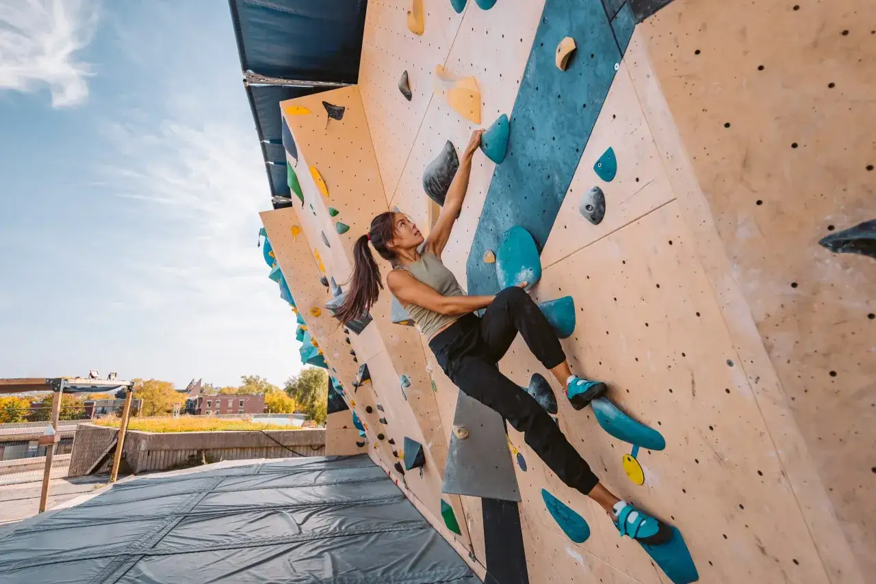 Frau beim training klettern an einer bunten Boulderwand im Freien. Sie greift nach einem blauen Griff, konzentriert und stark.