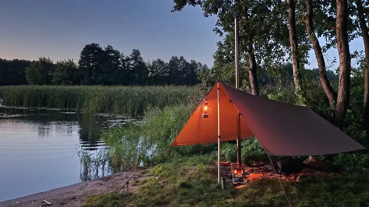 Ein gemütliches Lager am Wasser: Ein Tarp spendet Schutz, Laternen und ein kleiner Ofen sorgen für Licht und Wärme. Das ist Bushcraft – die Kunst des Überlebens in der Natur.