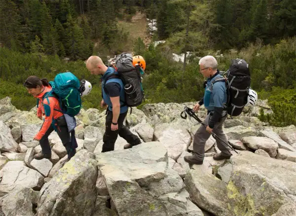 Drei Wanderer mit Rucksäcken und Helmen auf felsigem Untergrund. Die Ausrüstung auf den Rücken deutet auf eine Klettersteig-Packliste hin.