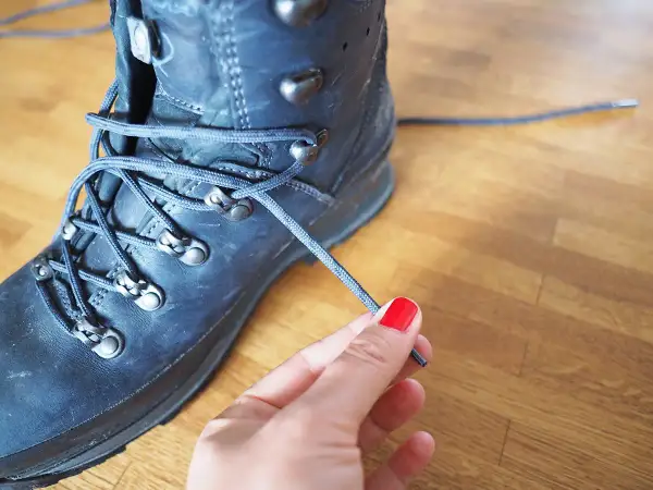 Eine Hand mit roten Nägeln bindet die Schnürsenkel eines robusten, dunklen Wanderschuhs. Bereit für das nächste Abenteuer.