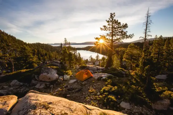 Ein orangefarbenes Zelt steht auf einem felsigen Hang mit Blick auf einen See, umgeben von Bäumen. Die Sonne bricht durch einen Baum und erzeugt einen Sterneneffekt. Ein Traum für jeden, der **wildcampen Europa** liebt.