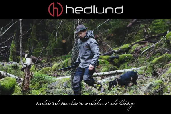 Ein Mann in Lodenkleidung und Hut geht mit einem Hund und einem Gewehr durch einen moosbewachsenen Wald.