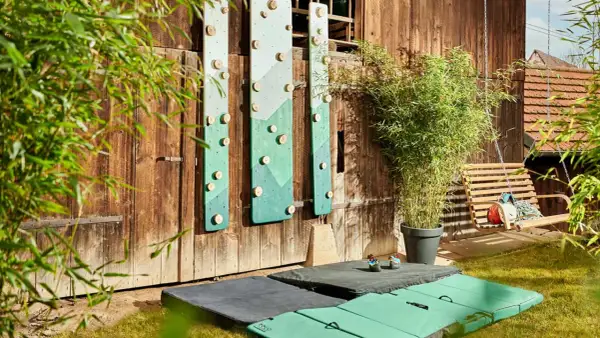 DIY-Kletterwand outdoor: Grüne Bretter mit Holzgriffen an Holzwand, daneben eine Schaukel und Klettermatten.