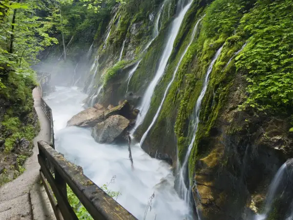 Ein Pfad führt entlang einer tosenden Klamm in Bayern, wo Wasserfälle über moosbewachsene Felsen stürzen.