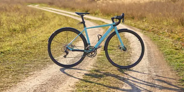 Ein hellblaues Fahrrad mit Schutzblechen steht auf einem Feldweg. Der Unterschied zwischen Cyclocross und Gravel wird hier deutlich: Dieses Rad ist bereit für Abenteuer auf Schotterwegen.