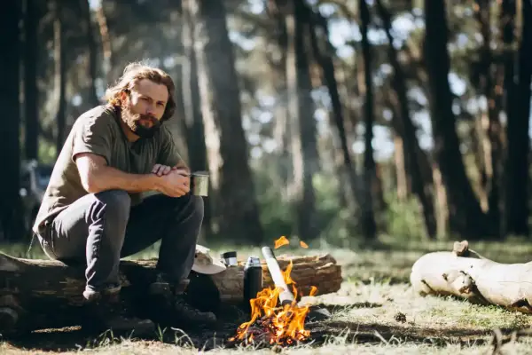 Mann sitzt am Lagerfeuer im Wald, hält eine Tasse. Das ist Bushcraft: Naturverbundenheit und Selbstversorgung.