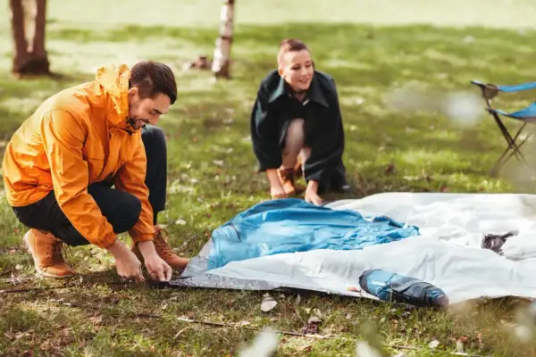 Mann und Frau bauen ein Zelt auf. Sie legen die Zeltplane aus und befestigen sie im Gras.
