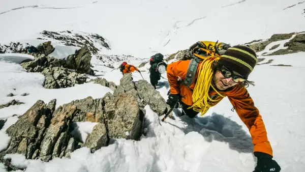Bergsteiger kämpfen sich durch den Schnee in den Berner Alpen. Ein Mann in Orange lächelt in die Kamera, während er sich an einem Felsen festhält.